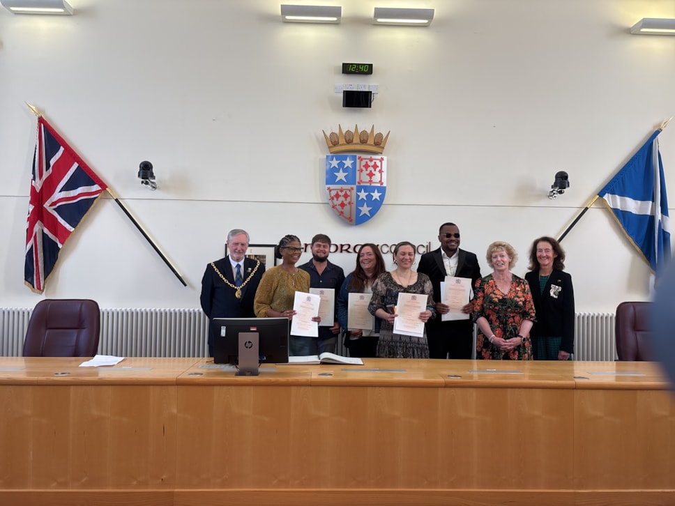 A group eight smiling people stand behind a long wooden desk. There's a coat of arms above them and a Union flag and Saltire at either side.