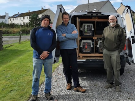 Three men stood in front of the back of a van containing animal cages