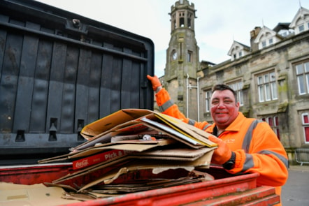 A man dressed in a hi-vis jacket holds open a bin filled with cardboard.