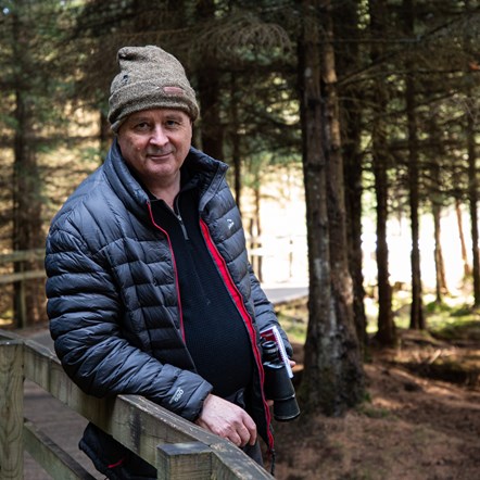 The Last Hillwalker author, John D. Burns, wears a navy waterproof insulated jacket and carries binoculars. Pine trees are in background.