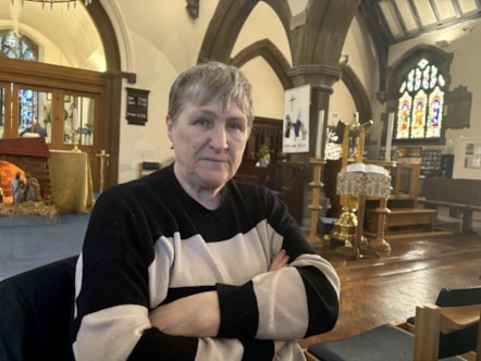 Sandra has short grey hair and is sat in the main area of the church wearing a black and white striped jumper looking into the camera