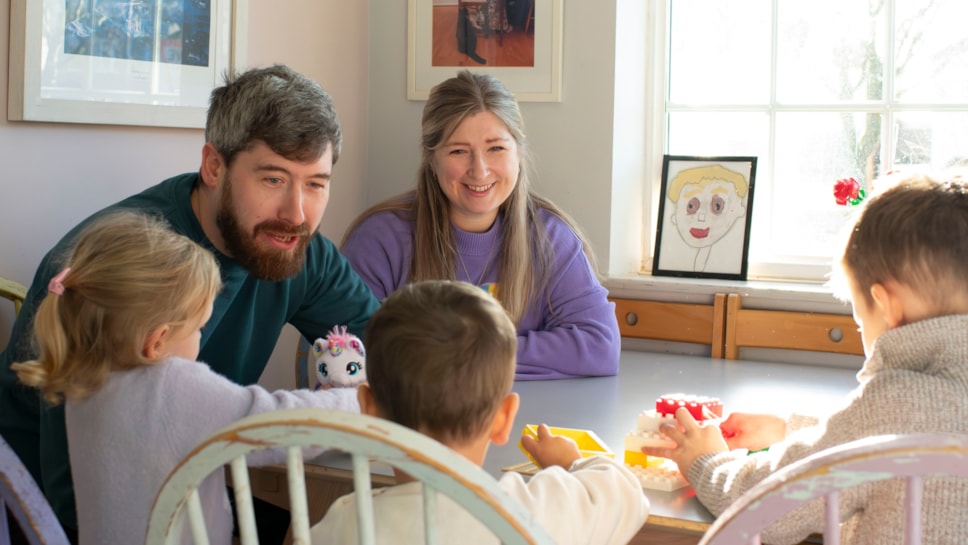 Paul and Rachael are sat at a table with three children. They are smiling and looking at the children