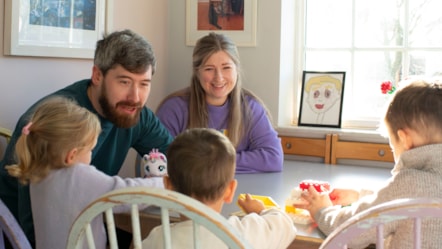 Paul and Rachael are sat at a table with three children. They are smiling and looking at the children