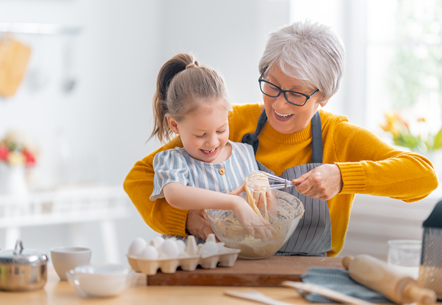 A kind foster carer and young girl laugh as they make a cake or dough. Child is around six and has her hair tied up, foster carer has short hair and a mustard jumper with an apron on. Positive, happy image.