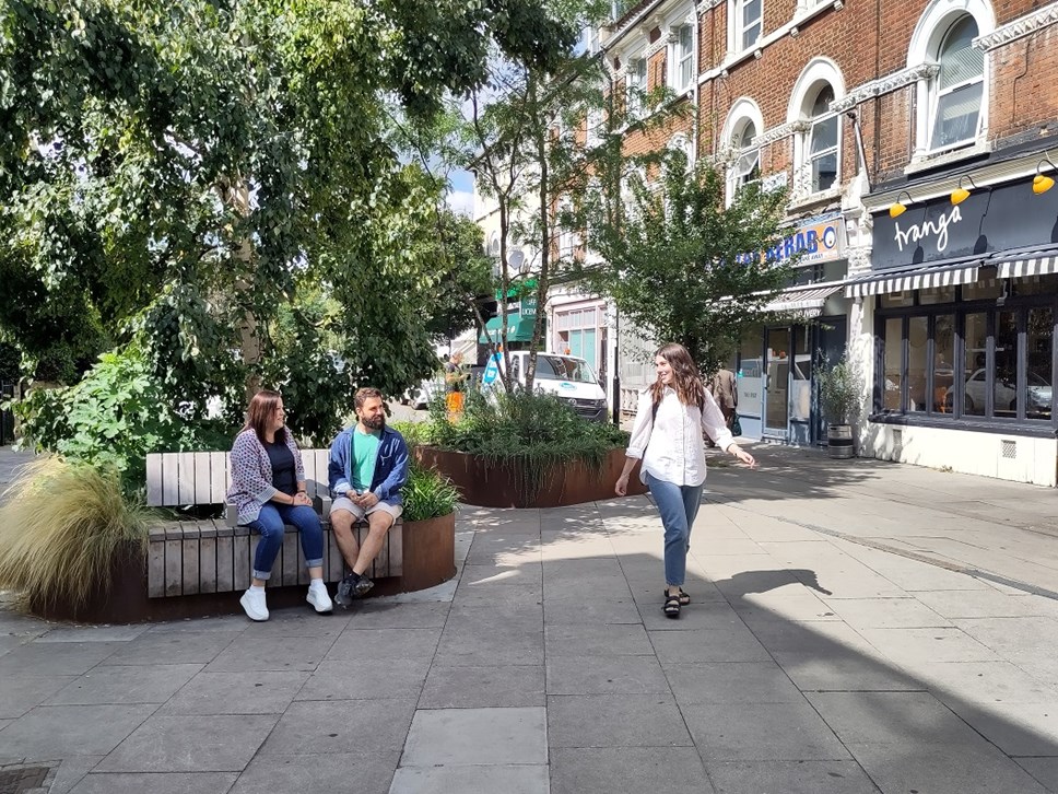 Local people enjoying a pocket park. Two people are sitting on a bench, and another is walking past.