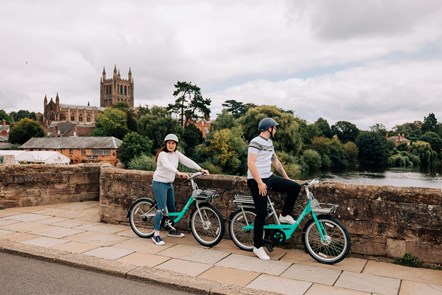 A lady and a man resting by a bridge overlooking a river in Hereford. The lady on the left of the picture is stood on a Beryl bike and the man on the right of the picture is stood on a Beryl e-bike.