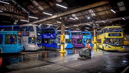 Old and new buses lined up in a depot, with a cleaner sweeping the floor in front of them.