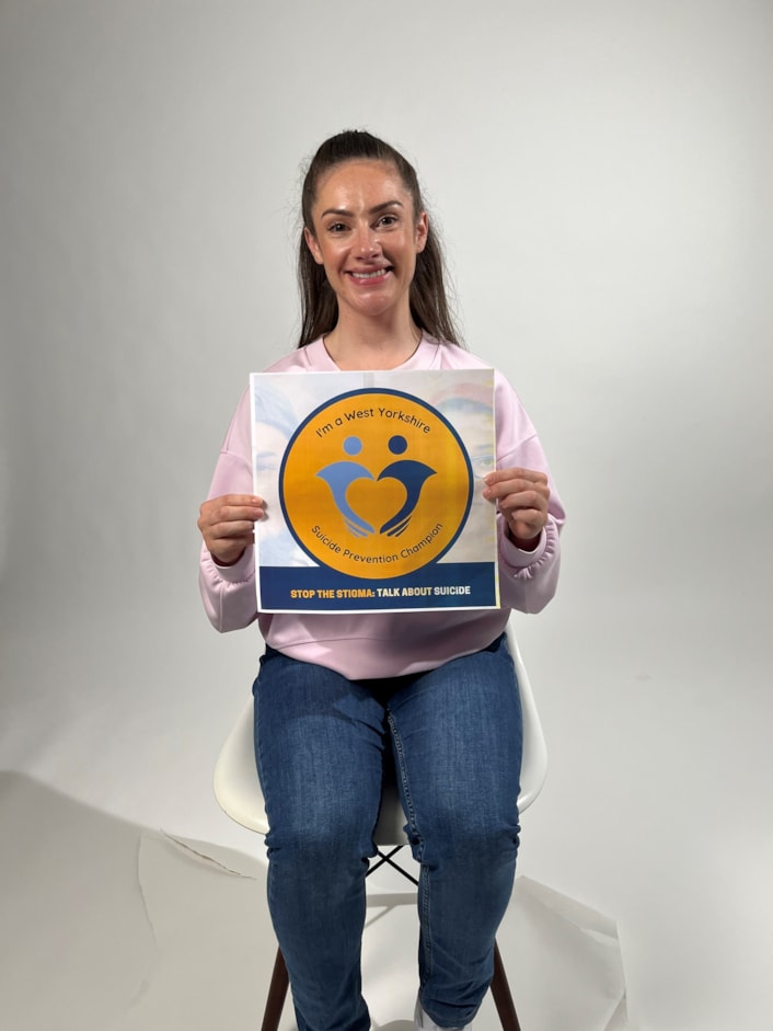 Suicide Prevention Champions - Isabel Hodgins: Isabel is pictured smiling at the camera, sitting on a chair, holding the Suicide Prevention Champion logo
