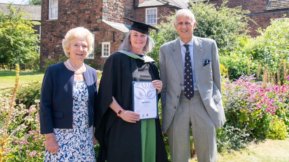 Three people one in centre wearing an academic gown and cap and holding certificate pictured in grounds of carlisle cathedral