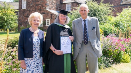 Three people one in centre wearing an academic gown and cap and holding certificate pictured in grounds of carlisle cathedral