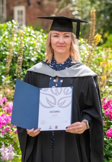 University of Cumbria graduate Lisa Scargill dressed in academic cap and robe holding a certificate stood in front of wildflowers